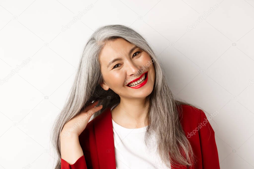 Beauty and aging concept. Close up of asian senior woman with red lips, long healthy grey hair, smiling at camera, standing over white background