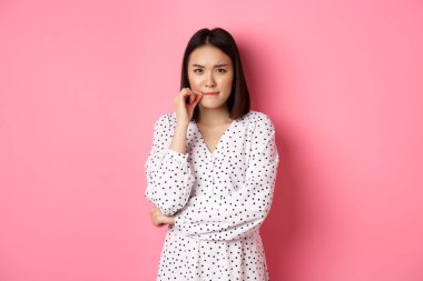 Esmer, Asyalı bir kadın hoşnutsuz görünüyor, kaşlarını çatıyor ve fermuarını çekiyor, dudakları umut dolu, pembe arka planda duruyor.
