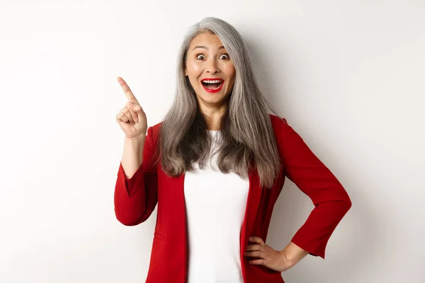 Affärsidé. Glada asiatiska affärskvinna med grått hår, bär röd blazer och makeup, pekar övre vänstra hörnet och ler förvånad, vit bakgrund — Stockfoto