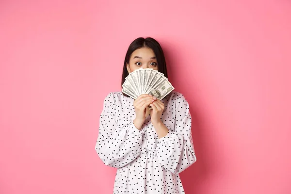 Concepto de compras. Linda mujer asiática escondiendo la cara detrás de dólares de dinero, mirando a la cámara, de pie sobre fondo rosa — Foto de Stock