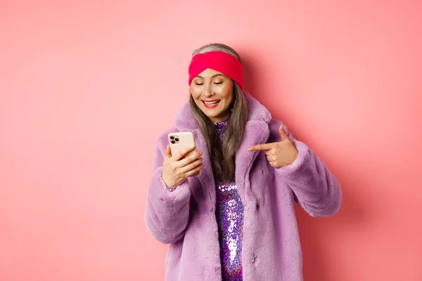 Online shopping and fashion concept. Cool asian senior woman checking out promotion on mobile phone, pointing finger at smartphone screen and smiling pleased, pink background — Stock Photo, Image