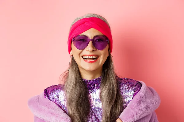 Mode. Nahaufnahme einer stilvollen Seniorin, die glücklich in die Kamera lächelt, Sonnenbrille und Disco-Stirnband trägt, rosa Hintergrund — Stockfoto