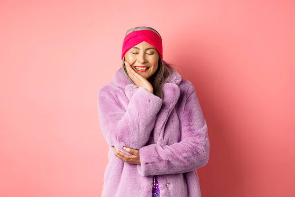 Moda e compras. feliz asiático maduro mulher no roxo falso casaco de pele sorrindo adorável e despreocupado, fechar os olhos com felicidade, de pé contra rosa fundo — Fotografia de Stock