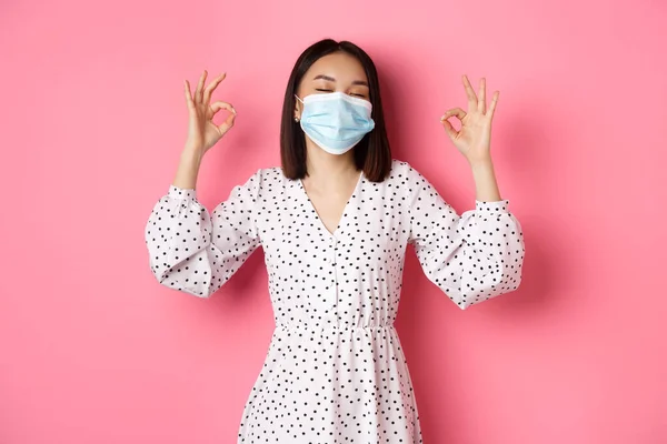Coronavirus, socialt distans och livsstil koncept. Nöjd asiatisk kvinna i ansiktsmask visar okej tecken, godkänna och samtycker, prisa bra val, rosa bakgrund — Stockfoto