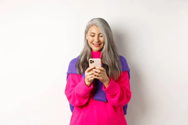 Piękna azjatycka seniorka w różowym, stylowym swetrze za pomocą smartfona. Starsza pani czyta przez telefon komórkowy i uśmiecha się, stojąc na białym tle — Zdjęcie stockowe