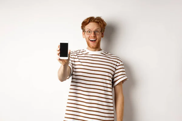 Concepto de tecnología y comercio electrónico. Feliz joven pelirrojo en gafas que muestran la pantalla del teléfono inteligente en blanco, mirando a la cámara sorprendida, de pie sobre fondo blanco — Foto de Stock