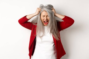 Distressed and pissed-off asian senior woman pulling hair and screaming, standing upset over white background