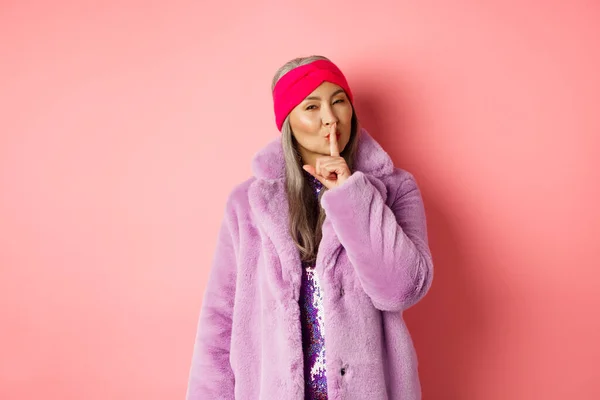 Concepto de moda y compras. Moderna mujer mayor china callando, contando un secreto y buscando astucia en la cámara, de pie en el abrigo de piel falsa de moda y diadema sobre fondo rosa — Foto de Stock