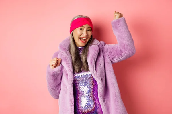 Einkaufskonzept. Fröhliche ältere asiatische Frau tanzt und hat Spaß, trägt eleganten lila Kunstpelz Mantel und Kleid, gewinnt den Preis und lächelt, steht über rosa Hintergrund — Stockfoto
