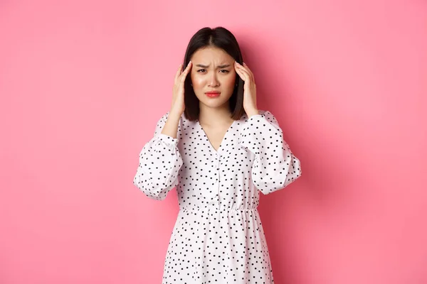 Imagen de mujer asiática disgustada con dolor de cabeza, malestar o mareo, ojos cerrados y cabeza masajeadora, migraña, de pie sobre fondo rosa —  Fotos de Stock