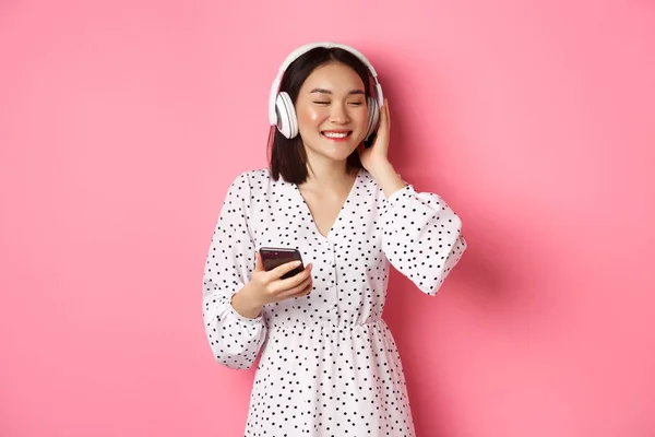 헤드폰으로 음악을 듣고, 눈을 감고 미소짓고, 휴대 전화를 들고, 핑크 색 배경 위에 서 있는 낭만적 인 아시아 소녀 — 스톡 사진