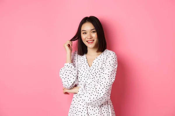 Bonito asiático feminino morena em vestido da moda, brincando com o cabelo e sorrindo flerte, de pé em vestido sobre fundo rosa — Fotografia de Stock