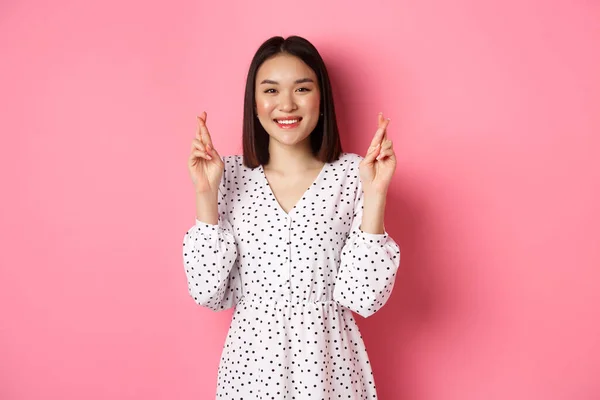 Bonito ásia mulher fazendo um desejo, sorrindo feliz e cruz dedos para boa sorte, esperando mais rosa fundo — Fotografia de Stock