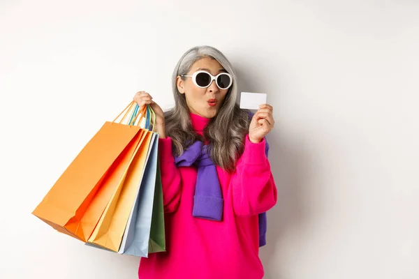 Moda asiática velha senhora vai às compras, segurando sacos de papel e cartão de crédito de plástico, vestindo óculos de sol elegantes, de pé sobre fundo branco — Fotografia de Stock