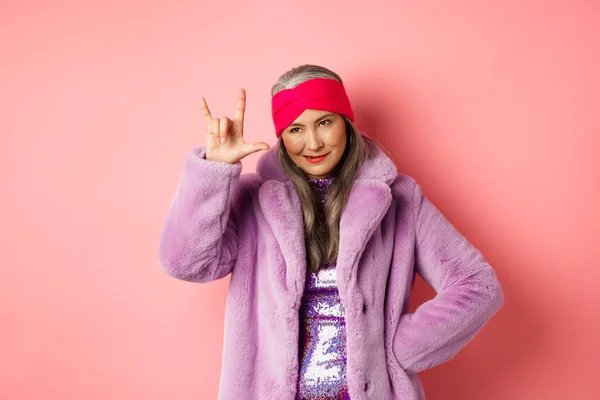 Engraçado e legal asiático sênior mulher mostrando rock n roll gesto, olhando atrevida, de pé sobre o fundo rosa — Fotografia de Stock
