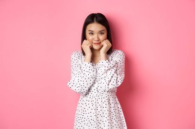 Tatlı ve utangaç Asyalı kız kızarıyor, yanaklarına dokunuyor ve kameraya aptalca bakıyor, pembe arka planda duruyor.