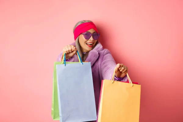 Funky och cool asiatisk senior kvinna i fashionabla kläder dans medan du går shopping på försäljning, hålla butik papperspåsar och ha kul, rosa bakgrund — Stockfoto