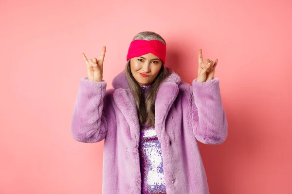 Cool asiático mulher madura na moda casaco de peles falso, mostrando sinais de chifres rock-n-roll e sorrindo atrevida para a câmera, se divertindo, de pé sobre fundo rosa — Fotografia de Stock