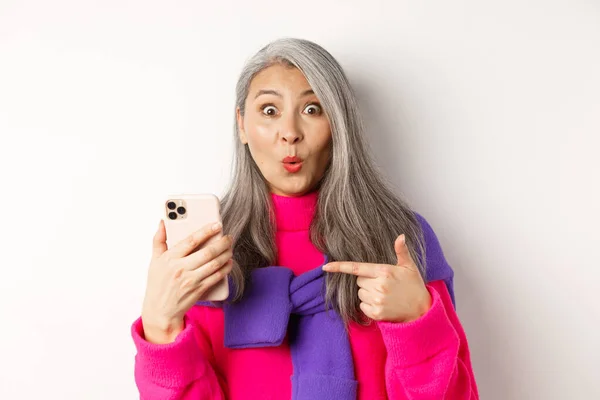 Achats en ligne. Gros plan de femme âgée asiatique étonnée pointant vers le smartphone et souriant étonné, debout sur fond blanc — Photo