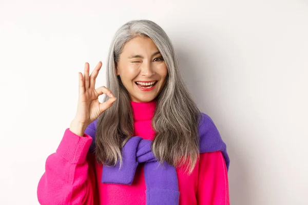 Primer plano de alegre abuela asiática en suéter rosa de moda, guiño y mostrando signo de bien, recomendando la promoción, de pie sobre fondo blanco — Foto de Stock