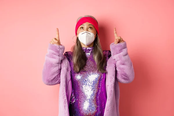 Coronavirus und Einkaufskonzept. Modische asiatische ältere Frau in Disco-Kleid und Atemschutzmaske schaut sich Promo-Angebot an, zeigt nach oben, rosa Hintergrund — Stockfoto