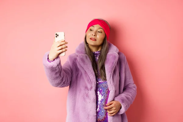 Modekonzept. Stilvolle asiatische Seniorin macht Selfie auf Smartphone, posiert in lila Kunstpelzmantel und Partykleid vor rosa Hintergrund — Stockfoto