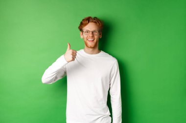 Kızıl saçlı yakışıklı genç adam, başparmağını kaldırıyor ve gülümsüyor memnun olmuş, iyi bir gözlük mağazasına övgüler yağdırıyor, yeşil arka planda duruyor.