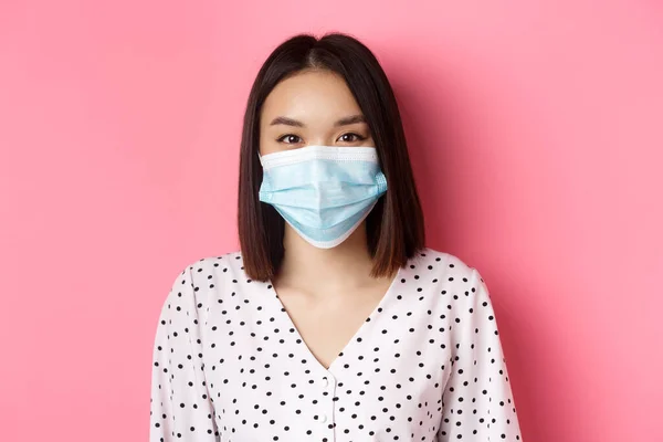 Covid-19, pandemi och livsstilskoncept. Närbild av vacker asiatisk kvinna i ansiktsmask ler med ögon, bär skydd mot coronavirus i offentliga, rosa bakgrund — Stockfoto