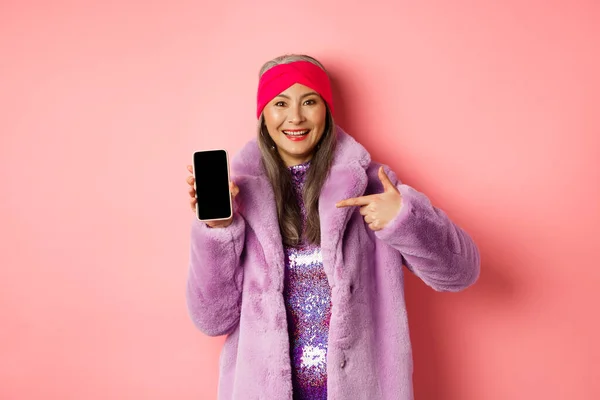 Online-Shopping und Modekonzept. Stilvolle asiatische reife Dame zeigt leeren Smartphone-Bildschirm, zeigt auf Telefon und lächelt, rosa Hintergrund — Stockfoto