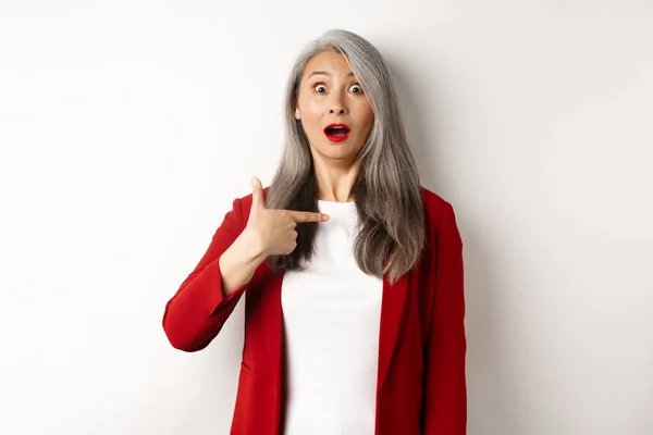 Förvånad asiatisk kvinna med grått hår, pekar på sig själv och kippar förvirrad, står över vit bakgrund — Stockfoto