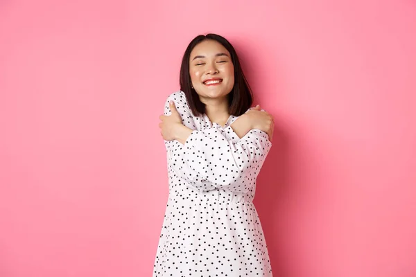 Mulher asiática bonita sentindo romântico, abraçando-se, fechar os olhos e sorrindo com rosto sonhador, de pé feliz sobre fundo rosa — Fotografia de Stock