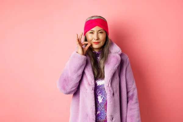 Мода и концепция шопинга. Хитрая азиатская старшая женщина в стильной одежде держит в секрете, щурясь и глядя коварно на камеру, молча рот, давая обещания, розовый фон — стоковое фото