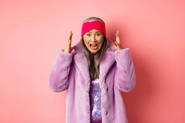 Ansioso asiático mulher sênior no elegante casaco de pele falso gritando, apertando as mãos e gritando alarmado e preocupado com a câmera, de pé sobre o fundo rosa — Fotografia de Stock
