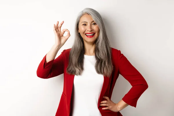 Asiatische Geschäftsfrau im roten Blazer, mit OK-Zeichen und lächelnd, zustimmend und wie etwas, vor weißem Hintergrund stehend — Stockfoto