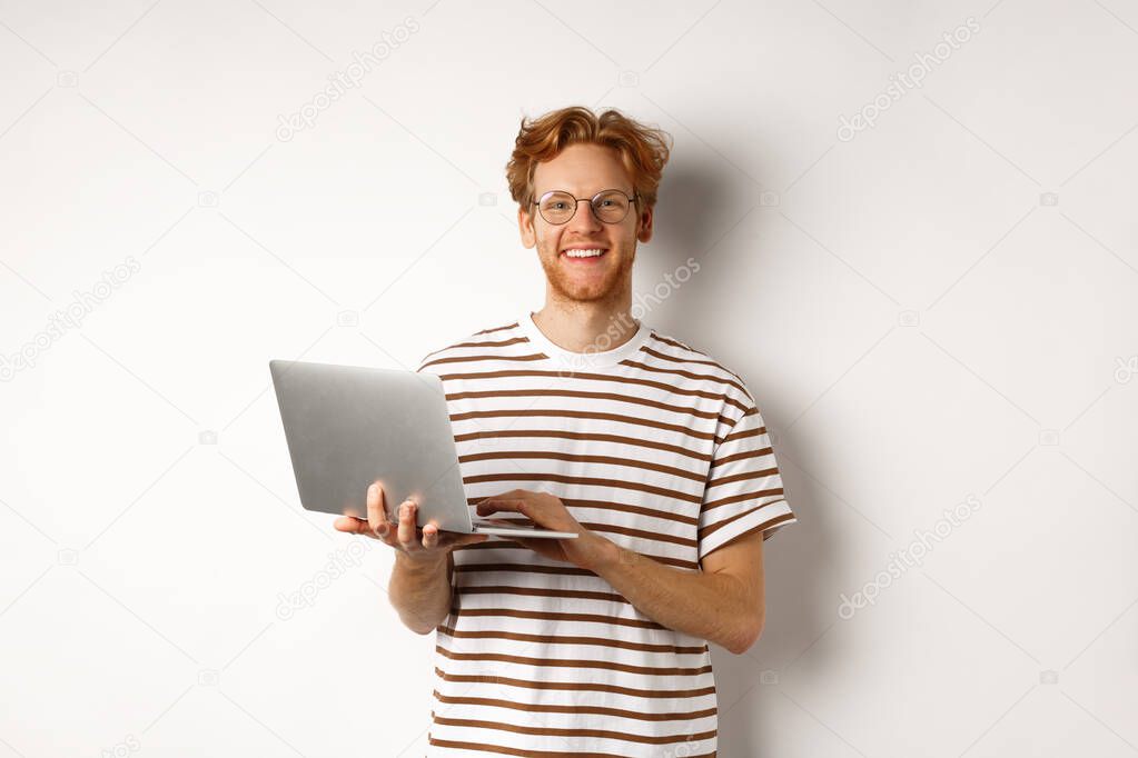 Smiling young man working on laptop and looking joyful, standing over white background