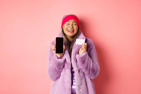 Besondere Förderung. Aufgeregte asiatische Seniorin zeigt leeren Smartphone-Bildschirm und Plastikkreditkarte, prüft Online-Angebot, rosa Hintergrund — Stockfoto