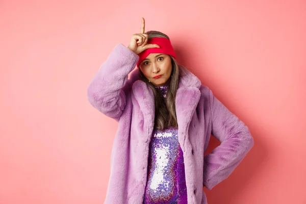 Stylish and cool asian mature woman showing loser sign on forehead, mocking you, standing in trendy faux fur coat and glittering dress, pink background — Stock Photo, Image