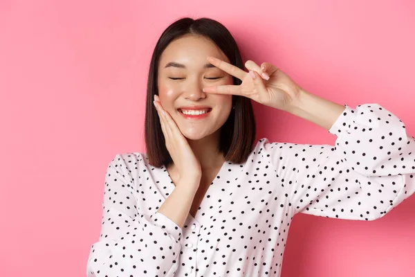 Concepto de belleza y estilo de vida. primer plano de bastante asiático mujer mostrando paz signo y tocando mejilla, sonriendo feliz a cámara, de pie sobre rosa fondo — Foto de Stock