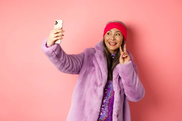 Modekonzept. Coole und gut gelaunte asiatische Seniorin macht Selfie mit Friedenszeichen, trägt trendigen Kunstpelzmantel und Partykleid, steht über rosa Hintergrund — Stockfoto