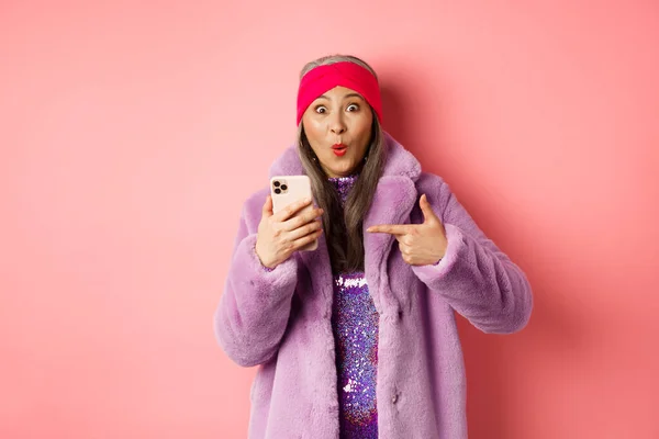 Online-Shopping und Modekonzept. Glückliche asiatische Großmutter checkt tolle Promo aus, zeigt mit dem Finger auf das Smartphone und schaut erstaunt, rosa Hintergrund — Stockfoto