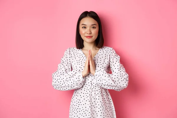 Hermosa señora asiática en vestido pidiendo ayuda, tomados de la mano en oración o gesto namaste, agradeciéndote, de pie sobre fondo rosa —  Fotos de Stock