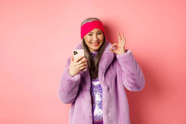 Online-Shopping und Modekonzept. Stilvolle asiatische Seniorin zeigt Okay Zeichen und hält Handy, empfiehlt Internet-Shop, rosa Hintergrund — Stockfoto