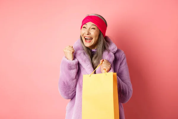 Shopping- und Modekonzept. Glückliche asiatische Seniorin gewinnt, hält Papiertüte in der Hand und macht Faust-Pump-Geste, sagt Ja mit fröhlichem Gesicht, steht über rosa Hintergrund — Stockfoto