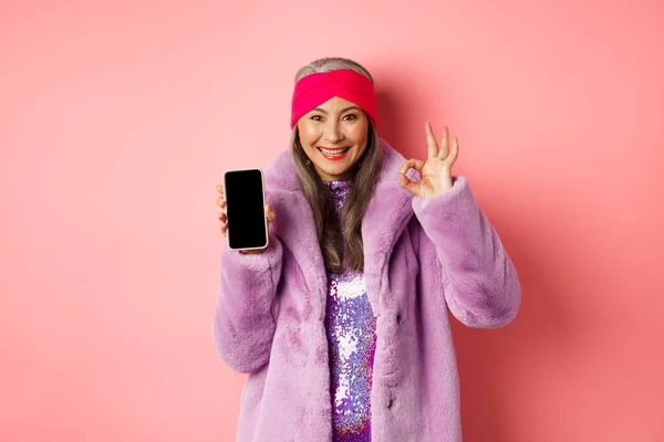 Conceito de compras online e moda. elegante sênior ásia mulher mostrando branco preto smartphone tela e OK sinal, demonstrar promoção, rosa fundo — Fotografia de Stock