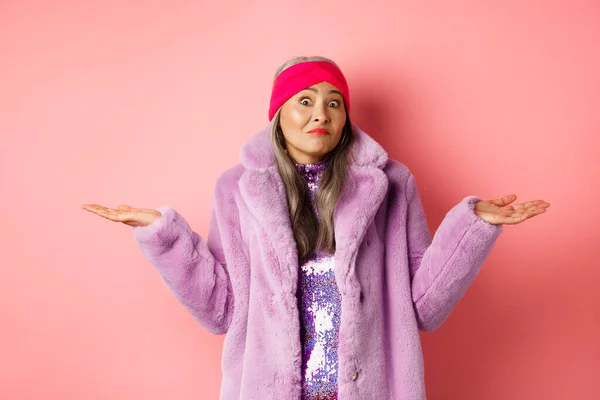 Модная и красивая старушка с опущенными плечами, выглядящая растерянной и неуверенной, стоящая в стильном фиолетовом пальто и головном уборе на розовом фоне — стоковое фото