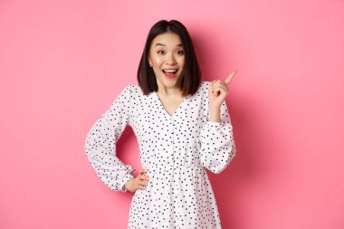 Heyecanlı güzel Asyalı kız sağ üst köşeyi işaret ediyor, şaşırmış ve kameraya bakıyor, mağaza tanıtım teklifini tartışıyor, pembe arka planda duruyor.
