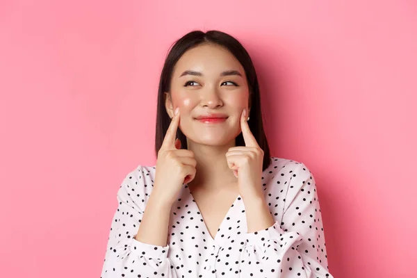 Conceito de beleza e cuidados com a pele. Close-up de bela mulher asiática cutucando bochechas e sorrindo satisfeito, de pé sobre fundo rosa — Fotografia de Stock