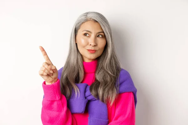 Primer plano de la mujer mayor coreana con estilo con el pelo gris mirando y señalando esquina superior izquierda, mostrando oferta de promoción, de pie sobre fondo blanco — Foto de Stock