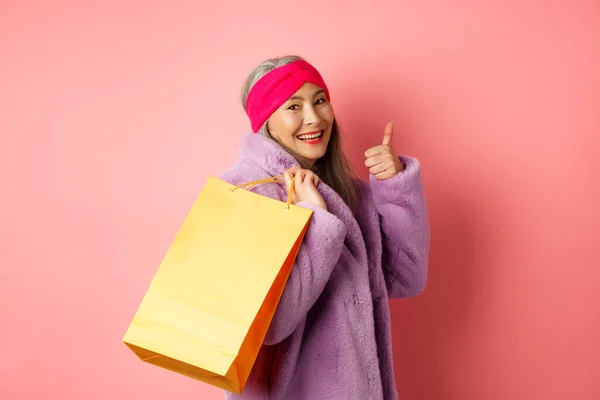 Stilvolle asiatische Seniorin geht einkaufen, trägt Papiertasche auf der Schulter und zeigt Daumen nach oben, empfiehlt Ladenrabatte, rosa Hintergrund — Stockfoto