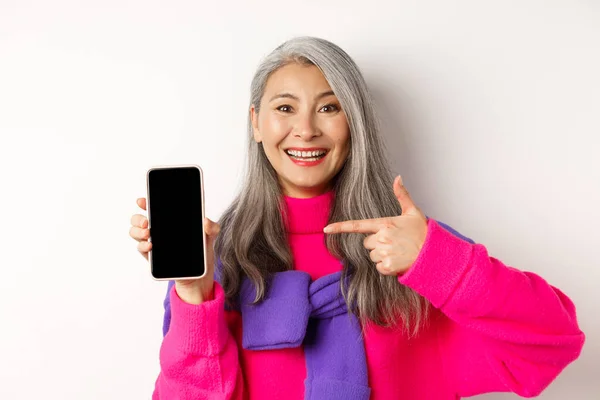 Shopping online. Bella nonna asiatica sorridente, indicando lo schermo vuoto dello smartphone, mostrando l'applicazione mobile, in piedi su sfondo bianco — Foto Stock